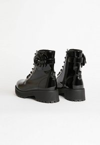 Botines negros brillantes con suelas gruesas, cremallera lateral, parte delantera con cordones y una gran flor decorativa en el tobillo exterior.