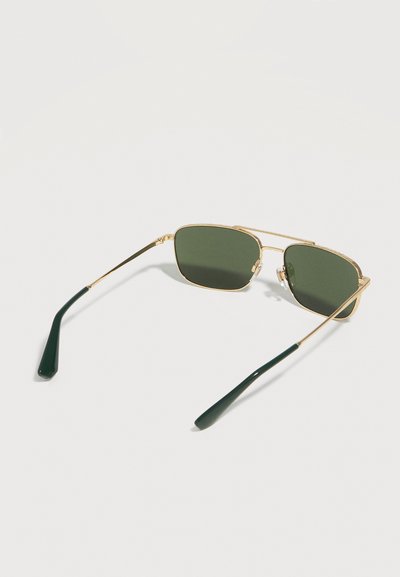 Ορθογώνια γυαλιά ηλίου με χρυσή μεταλλική κορνίζα, σκούρους πράσινους φακούς και μαύρες άκρες πλαισίων που ακουμπούν σε λευκή επιφάνεια.