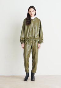 Hoodie od umjetne kože u maslinasto zelenoj boji i usklađene hlače s elastičnim pojasom, manžetama i vezicom. Glatka tekstura, minimalistički dizajn. Crne gležnjače.