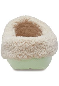 Zapatilla de felpa beige con un upper suave y aterciopelado y una suela de goma texturizada de color verde claro. El diseño presenta una forma redondeada y una abertura amplia.