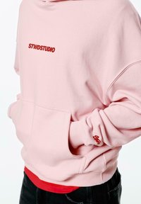 Light pink hoodie with front pocket, ribbed cuffs, and embroidered red text "STWDSTUDIO" on the chest, layered over a red shirt.