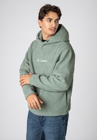 Groene, gestructureerde fleece hoodie met een kangoeroezak. Voorzien van een trekkoord bij de capuchon en een wit logo op de voorkant. Gepaard met blauwe spijkerbroek.