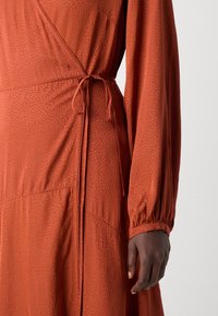 Robe portefeuille à manches longues de couleur rouille avec un motif à pois subtil, nouée à la taille, portée par une personne à la peau foncée.