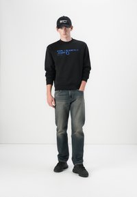 Sudadera negra con logo azul, jeans de mezclilla grises y zapatillas negras. El modelo lleva una gorra negra y está de pie contra un fondo blanco.