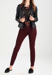 Zwarte leren jas met geribbelde schouderdetails, top met bloemenpatroon, bordeauxrode strakke broek en zwarte instapschoenen met vachtaccenten.