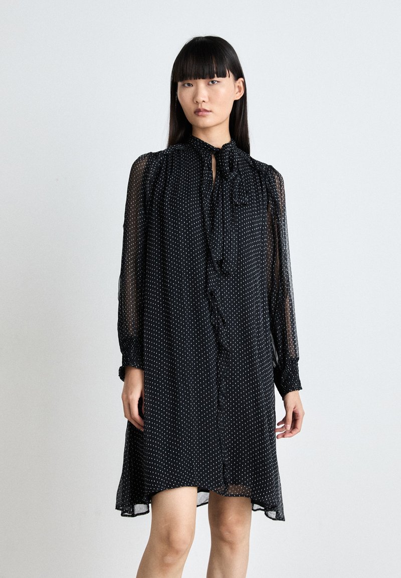 Schwarzes Kleid mit transparenten langen Ärmeln, gefälteltem Ausschnitt und Bindeband-Detail. Verziert mit einem weißen Punktmuster und einer durchgehenden Knopfleiste.