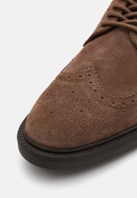 Scarpa in camoscio marrone con dettagli perforati intricati, punta arrotondata e suola in gomma scura. Il primo piano mette in risalto la texture e gli elementi di design.