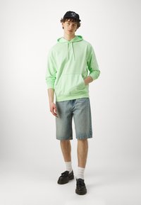 Sweatshirt verde claro com cordão e bolso frontal, combinado com calções de ganga azuis, meias brancas e sapatos pretos. Design simples e ajuste descontraído.