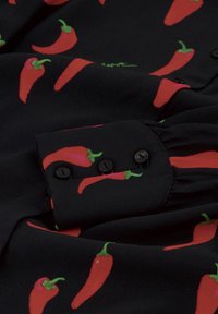 Black fabric blouse with a red chili pepper pattern. Features a buttoned cuff with black buttons and gathered detail on the sleeve.