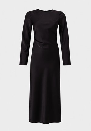 Femme portant une longue robe noire en satin à manches longues et des talons hauts noirs à brides, debout devant un fond clair uni.