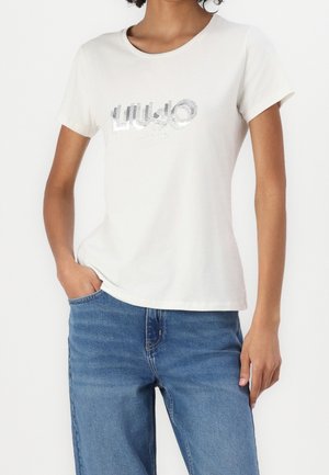 Frau trägt ein weißes kurzärmeliges T-Shirt mit silbernen Pailletten-Schriftzug und blaue Jeans, steht mit einer Hand in der Hosentasche.