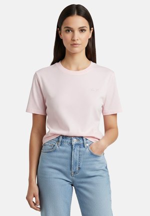 Donna con capelli lunghi e scuri che indossa una T-shirt a maniche corte rosa chiaro infilata in un paio di jeans chiari a vita alta, in piedi con una mano nella tasca.