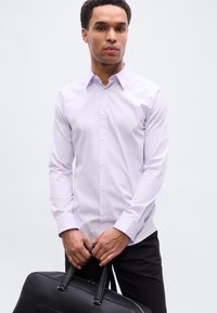 Ανοιχτό μωβ πουκάμισο με μακριά μανίκια, γιακά, κούμπωμα με κουμπιά και μία τσέπη. Το μοντέλο κρατάει μαύρη δερμάτινη τσάντα. Ουδέτερο φόντο.