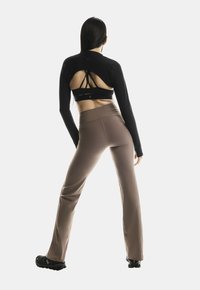 Žena stojí so šírkou nôh, oblečená v čiernom dlhom rukáve crop top s výrezom na chrbte, športovou podprsenkou s popruhmi, hnedých legínach a čiernych športových topánkach.
