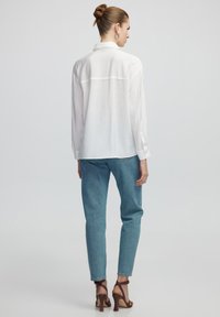 Camisa blanca de manga larga con un ajuste relajado, que presenta una costura en el yugo trasero, combinada con jeans ajustados de color azul claro y sandalias de tacón marrones.