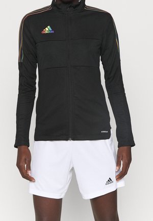 Persoon draagt een zwarte Adidas jas met rits en regenbooglogo en witte Adidas shorts met zwart logo, staand tegen een effen achtergrond.