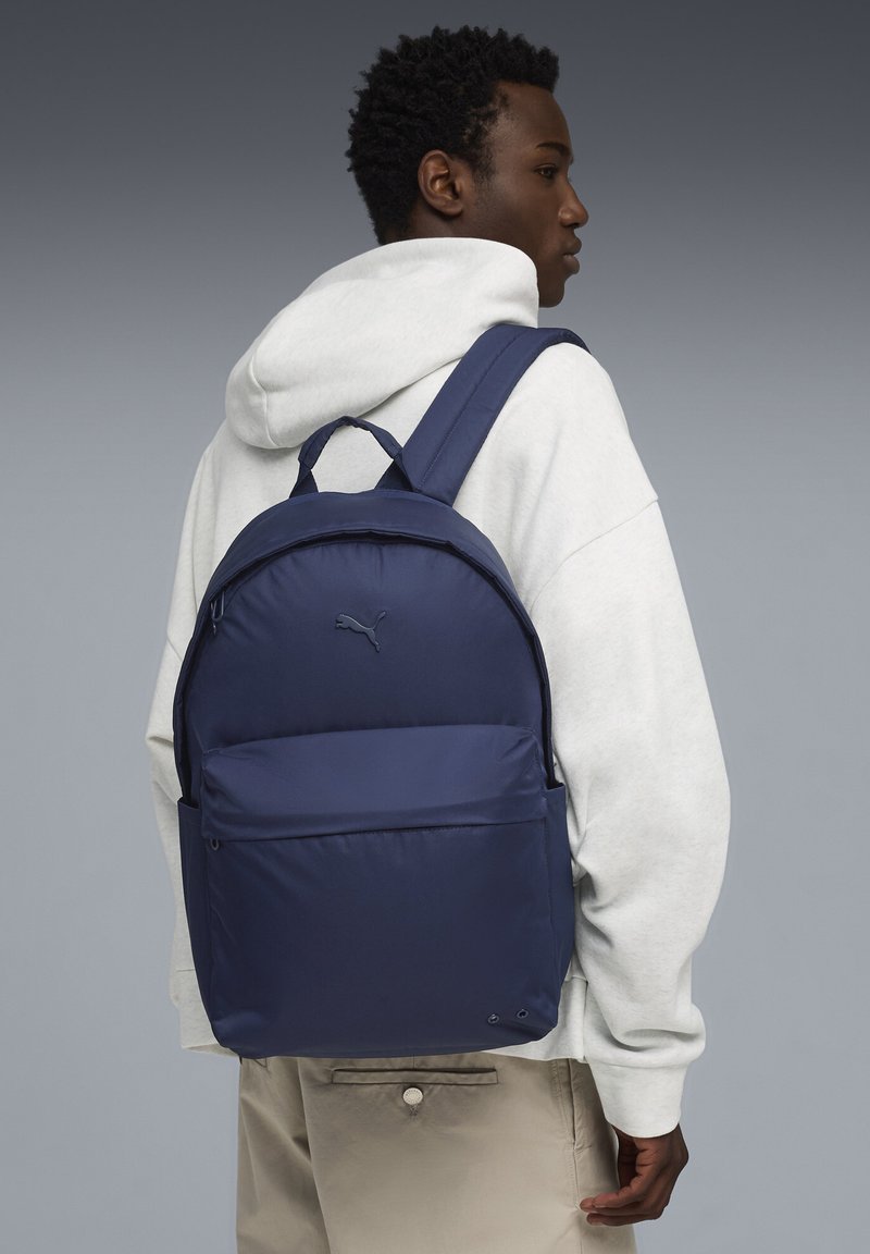 Sac à dos bleu marine avec dos rembourré, bretelles ajustables, poche avant et logo Puma. Matériau lisse et durable, conçu pour le confort.