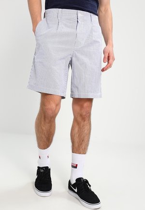 Homme portant un short rayé gris clair, des chaussettes blanches et des baskets noires, debout avec les mains partiellement dans les poches.