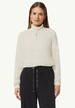 Weißer strukturierter Pullover mit hohem Kragen und halbem Reißverschluss, kombiniert mit hochgeschnittenen schwarzen Hosen mit einem gebundenen Gürtel.