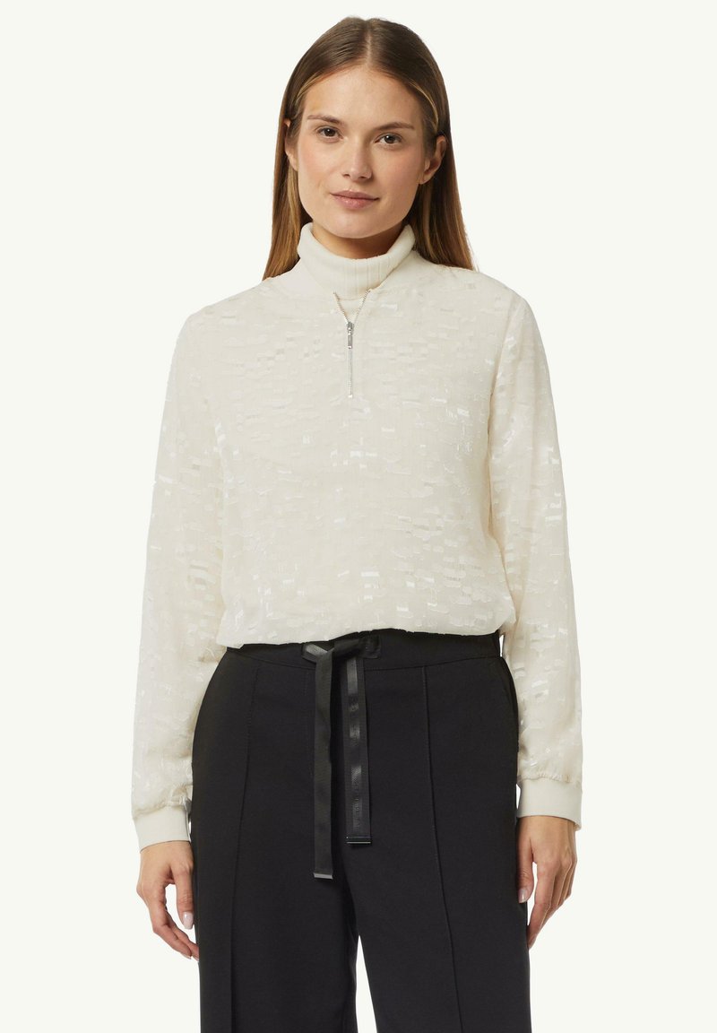 Weißer strukturierter Pullover mit hohem Kragen und halbem Reißverschluss, kombiniert mit hochgeschnittenen schwarzen Hosen mit einem gebundenen Gürtel.