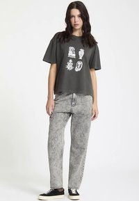 Donkergrijs oversized T-shirt met witte grafische prints, gecombineerd met vervaagde grijze jeans met rechte pijpen en zwarte sneakers met crèmekleurige accenten.
