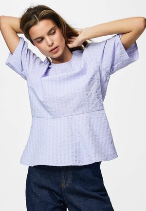 Młoda kobieta poprawia włosy, nosząc jasnofioletowy teksturowany krótki top z krótkim rękawem i ciemnoniebieskie dżinsy na jednolitym białym tle.