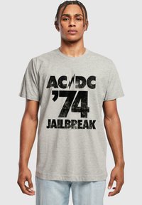 Pilkas medvilninis marškinėlis su ryškiu juodu grafiniu piešiniu, kuriame užrašas "AC/DC '74 JAILBREAK" ir žaibo dizainas, apvalia iškirpte ir trumpomis rankovėmis.