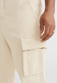 Beige corduroy cargo-housut, joissa on uritettu pinta, sivutasku ja huomaamaton logo-ominaisuus. Kangas näyttää pehmeältä ja kestävältä.