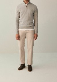 Helles grauer Reißverschluss-Pullover, darunter ein cremefarbenes Hemd, beiges Chino, und dunkelbraune Wildlederstiefel. Weiche Textur und lässige Passform.