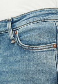 Jeans in denim blu con tasca frontale con cuciture arancioni, dettagli in rivetti metallici e una texture leggermente sbiadita.