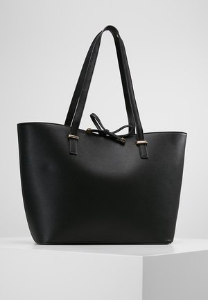 Schwarze Ledertasche mit doppelten Schulterriemen und einer Schleifendetail vorne, präsentiert auf einem weißen Podest vor einem grauen Hintergrund.