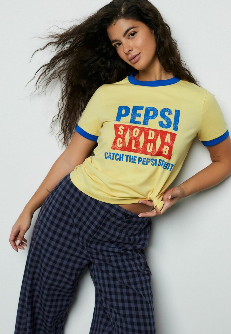 Maglietta gialla a maniche corte con colletto a contrasto blu, con la grafica "PEPSI SODA CLUB" in blu e rosso; abbinata a pantaloni a quadri neri e blu.