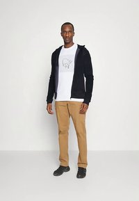 Felpa nera con zip sopra una t-shirt bianca con grafica, abbinata a pantaloni cargo beige e scarpe nere. Il modello è in piedi contro uno sfondo neutro.