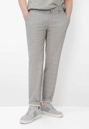 Uomo che indossa pantaloni a quadri grigio chiaro con risvolti e sneakers abbinate grigio chiaro, in piedi con le mani nelle tasche.