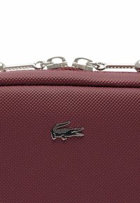 Sac en simili cuir texturé bordeaux avec des accessoires en métal argenté et un logo crocodile noir à l'avant. Fermeture zippée sur le dessus.
