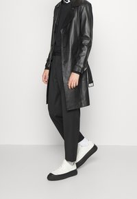 Schwarzer Ledertrenchcoat mit großem Kragen, doppelt geknöpften Knöpfen und verstellbaren Manschetten, kombiniert mit schwarzen Hosen und weißen Sneakers.