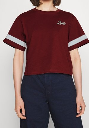 Osoba nosząca bordowy krótki top z szarymi paskami na rękawach i logo Levi's, w połączeniu z ciemnoniebieskimi spodniami z wysokim stanem.