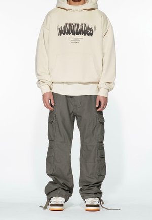 Person trägt beige Kapuzenpullover mit schwarzem Grafiktext, lockere graue Cargohosen mit mehreren Taschen und weiße Sneaker mit orangenen Sohlen.
