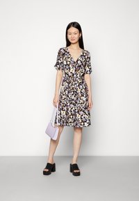Vestido floral em castanho escuro com mangas curtas, cintura atada e saia até ao joelho. Combinado com sapatos pretos de salto bloco e uma mala de cor lilás clara.