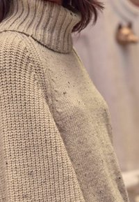 Gros plan sur une personne portant un pull à col roulé en maille épaisse beige avec des coutures texturées et des détails de fil moucheté.