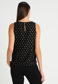 Haut noir sans manches avec une ouverture en forme de goutte d'eau dans le dos, orné de pois métalliques dorés disposés de manière uniforme sur le tissu.