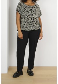 Frau, die schwarze Hosen, schwarze Loafers mit goldenen Details und ein kurzärmeliges Oberteil in Schwarz-Beige-Muster trägt und auf einem Holzboden steht.