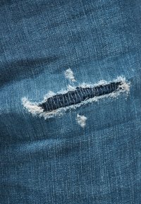 Primer plano de tela vaquera azul con un desgarro horizontal que muestra bordes blancos deshilachados y tela más oscura debajo.
