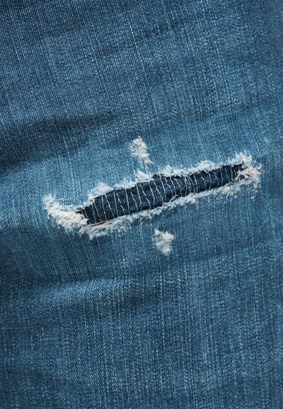 Κοντινή λήψη μπλε υφάσματος ντενίμ με οριζόντια σχισμή που δείχνει άσπρες φθαρμένες άκρες και πιο σκούρο ύφασμα από κάτω.