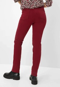 Jeans elasticizzati color bordeaux con vestibilità slim, dotati di due tasche posteriori e una leggera svasatura alla caviglia, abbinati a stivaletti neri su uno sfondo bianco.