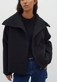 Cappotto in misto lana nero con ampio colletto, chiusure a bottone e una vestibilità comoda. Indossato sopra un maglione in maglia nera e pantaloni in denim blu.