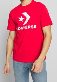 Punainen puuvillatee-paita, jossa on suuri valkoinen tähti ja nuoli -grafiikka, sekä "CONVERSE" tulostettuna selkeästi alle valkoisena. Lyhyet hihat, pyöreä pääntie.