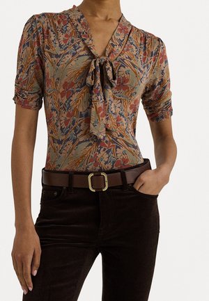 Kobieta nosząca bluzkę w kwiatowy wzór z wiązaniem przy szyi, w połączeniu z ciemnobrązowymi spodniami z wysokim stanem i pasem w kolorze brązowym z złotą klamrą.