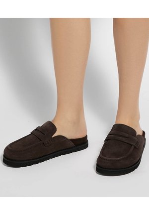 Personne portant des mocassins marron foncé sans lacets avec des semelles noires, montrée de la moitié du mollet vers le bas sur un fond uni.