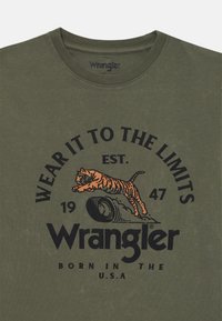 T-shirt in cotone verde oliva con un design grafico nero e arancione di una tigre in salto e il testo "Indossalo fino ai limiti" e "Wrangler."